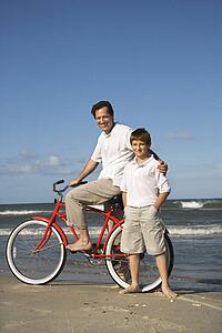 Caucasian father and pre-teen boy standing on beach with bicycle.