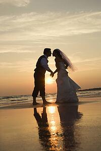 Caucasian prime adult male groom and female bride holding hands and kissing barefoot on beach at sunset.