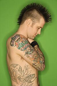 Profile of Caucasian man with mohawk and tattoos standing against green background.