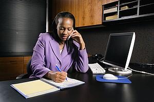 African-American young adult business woman talking on cell phone and writing in planner in office.