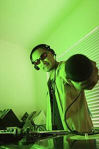 Asian young adult male DJ standing behind mixing equipment and turntable holding microphone out to viewer.