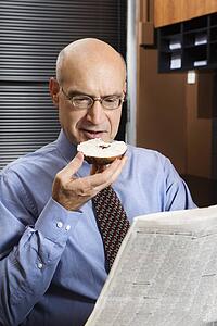 Caucasian middle-aged businessman sitting in office reading newspaper while eating a bagel with cream cheese.