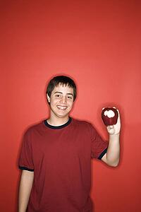 Portrait of smiling Caucasian teen boy holding partially eaten apple against red background.