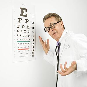 Mid-adult Caucasian male doctor wearing eyeglasses making gesture with eye chart in background.