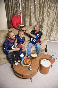 Mother serving snacks to male African-American family members during football game. 