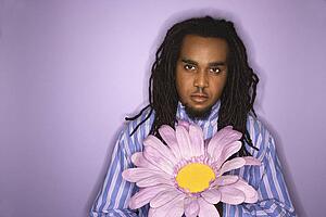Serious African-American mid-adult man holding big fake flower on purple background.