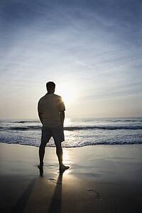 Caucasian mid-adult man standing alone on beach looking at ocean at sunrise.