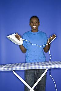 Portrait of smiling African-American teen boy holding iron and cord standing in behind ironing board against blue background.