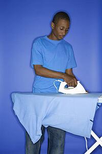 Portrait of African-American teen boy ironing shirt standing against blue background.