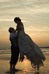 Caucasian mid-adult groom lifting up bride on beach at sunset.