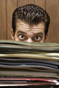 Caucasion mid-adult retro businessman peering with wide eyes over a tall stack of folders.