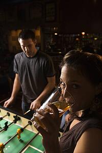 Young woman drinking beer and looking at viewer while man plays foosball in pub.