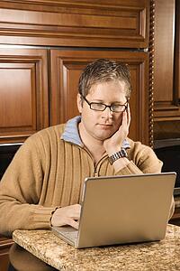 Caucasian man working on laptop computer in kitchen.