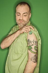 Portrait of Caucasian man pulling up sleeve to show tattoo standing against green background.