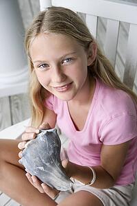 Caucasian pre-teen girl sitting indian style in rocking chair holding conch shell looking at viewer and smiling.