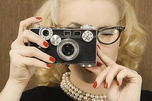 Mid-adult Caucasian female in vintage outfit holding a vintage film camera up to her face.