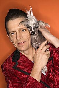 Caucasian mid-adult male wearing velvet jacket and holding Chinese Crested dog.