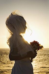 Young adult female Caucasian bride at sunset holding bouquet on beach.