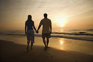 Caucasian mid-adult couple walking holding hands on beach at sunset.
