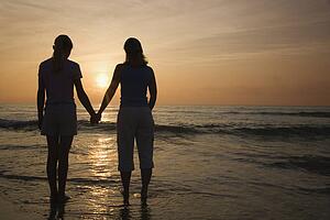 Caucasian mid-adult mother teenage and daughter standing on beach at sunset holding hands.
