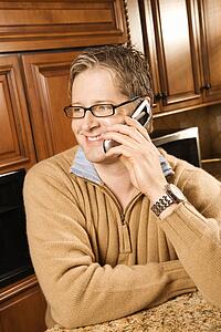 Caucasian man smiling and talking on cellphone while leaning on kitchen counter.