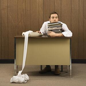 Mid-adult Caucasian male holding stack of paperwork with hand on calculator with long printout.