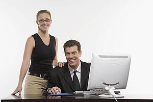 Caucasian mid-adult woman touching shoulder of man sitting at computer and looking at viewer.