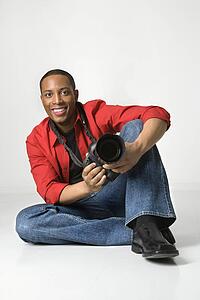African American young male adult sitting on floor with camera looking at viewer smiling.
