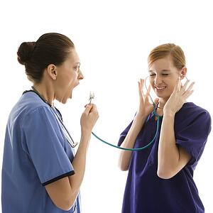 Portrait of Caucasian healthcare workers wearing scrubs with one yelling into stethoscope that is attached to other's ears against white background.