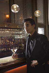 Prime adult Asian male standing at bar with cocktail.