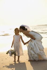 Caucasian mid-adult bride kneeling to give flower girl a kiss on the cheek while holding hands barefoot on beach.