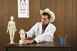 Mid-adult Caucasian male doctor wearing cowboy hat sitting at desk playing with figurine.
