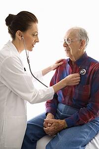 Mid-adult Caucasian female doctor listening to elderly Caucasian male's heart with stethoscope.