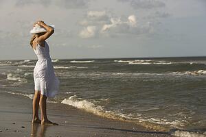 Caucasian pre-teen girl standing on beach holding hat on head.