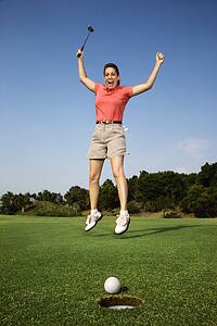 Caucasion mid-adult woman holding golf club jumping in air cheering with golf ball and hole in foreground.