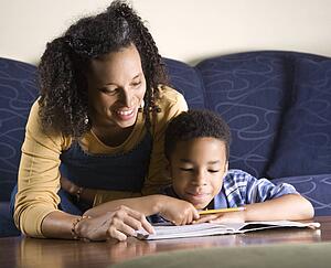 Portrait of young boy doing homework while mother looks over shouulder.