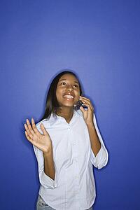 Portrait of smiling African-American teen girl looking up and talking on cellphone standing against blue background.