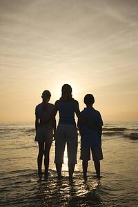 Caucasian mid-adult mother and teenage kids standing silhouetted on beach at sunset.