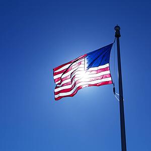 American flag on flagpole waving against blue sky with sun shining through from behind.