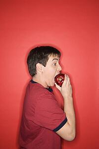 Portrait of Caucasian teen boy biting an apple standing against red background.