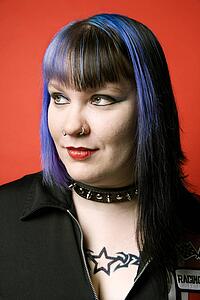 Portrait of Caucasian woman with blue hair, tattoo, and spike collar against orange background.