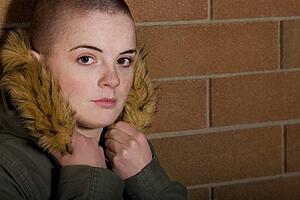 Teen girl with shaved head holding onto hood of coat and leaning on brick wall