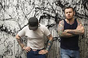 Two Caucasian tattooed men standing against paint splattered background.