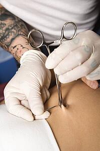 Skin held in piercing tool as needle is ready to go in to pierce naval
