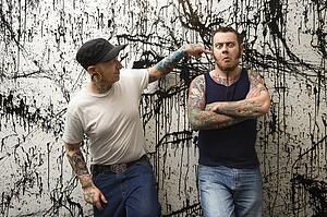 Two Caucasian tattooed men standing against paint splattered background.
