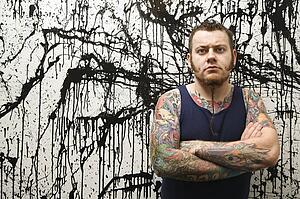 Tattooed man with braided beard standing against paint splattered background
