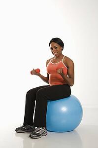 Smiling African American young adult woman sitting on exercise ball holding dumbbells.