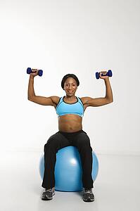 African American young adult woman sitting on exercise ball and raising dumbbells over head.