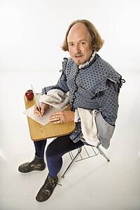 William Shakespeare in period clothing sitting in school desk writing with quill pen.