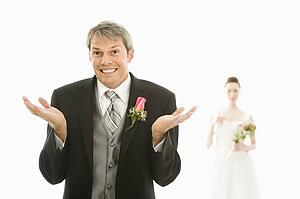 Groom in foreground with hands raised in air and bride in background